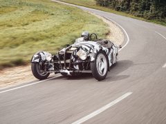 Een Morgan 3 Wheeler met een geometrisch camouflagepatroon rijdt op een bochtige weg, omgeven door gras en bomen. De bestuurder draagt een helm.