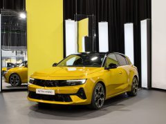 Een gele Opel Astra Hybrid-auto wordt tentoongesteld in een showroom met heldere verticale lichten op de achtergrond en een spiegel die een deel van het voertuig weerspiegelt.