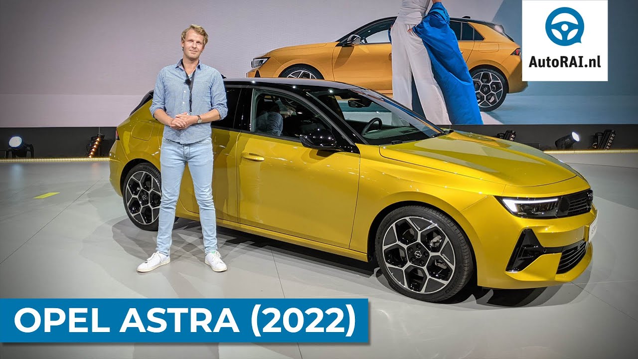 Een persoon staat naast een felgele Opel Astra (2022) op een podium. Het logo ‘AutoRAI.nl’ en de tekst ‘OPEL ASTRA (2022)’ zijn prominent weergegeven. Op de achtergrond wordt op een groot scherm nog een afbeelding van de gestroomlijnde Opel Astra getoond.