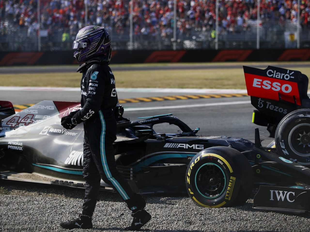 Een autocoureur loopt weg van een stilstaande Mercedes-AMG Petronas Formule 1-auto op een grindterrein, met toeschouwers op de tribunes op de achtergrond. De vastberaden uitdrukking van Max Verstappen weerspiegelt de intense sfeer van het racecircuit.