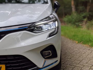 Close-up van de koplamp rechtsvoor van een Renault Clio E-Tech Hybrid met verlicht LED-licht, geparkeerd op een geplaveid pad in een bosrijke omgeving.
