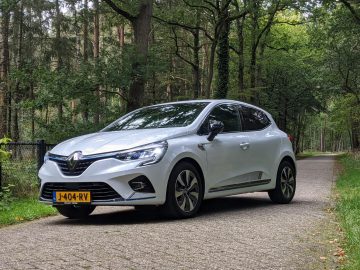 Op een verhard pad in een bosrijke omgeving staat een witte Renault Clio E-Tech Hybrid met Nederlands kenteken geparkeerd.