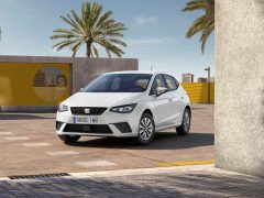 Op een zonnige dag staat een witte Seat Ibiza buiten geparkeerd, vlakbij een geel en grijs geometrisch bouwwerk met palmbomen en moderne gebouwen op de achtergrond.