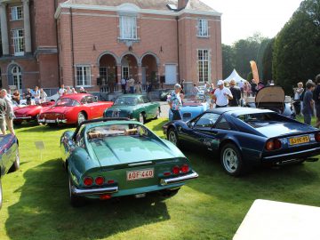 Een groep mensen bekijkt klassieke auto's tentoongesteld op een grasveld voor een groot bakstenen gebouw op een zonnige dag tijdens het Antwerp Concours 2021.
