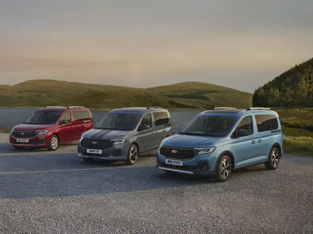 Drie veelkleurige minibusjes, waaronder een Ford Tourneo Connect, staan geparkeerd op een grindoppervlak nabij een meer met heuvels zichtbaar op de achtergrond. Het rode busje bevindt zich aan de linkerkant, het grijze busje in het midden en het blauwe busje aan de rechterkant.