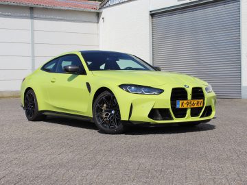 Op een verhard oppervlak op een industrieterrein staat een knalgele BMW M4 Competition Coupé geparkeerd, met op de achtergrond een roldeur en een gebouw. Kentekennummer K-956-RV.