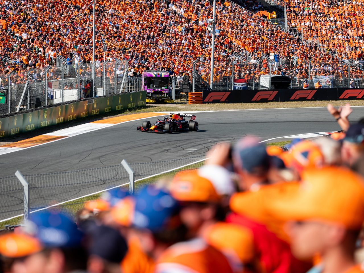 Een Formule 1-auto rijdt een bocht op een racecircuit terwijl een grote menigte toeschouwers, waarvan velen oranje gekleed zijn, toekijken vanaf de tribunes en de zijlijn.