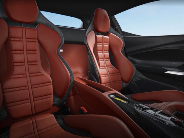 De afbeelding toont het interieur van een Ferrari 296 GTB met twee roodleren kuipstoelen, een middenconsole met bedieningselementen en een modern dashboard. De ramen van de auto laten buiten een helderblauwe lucht zien.