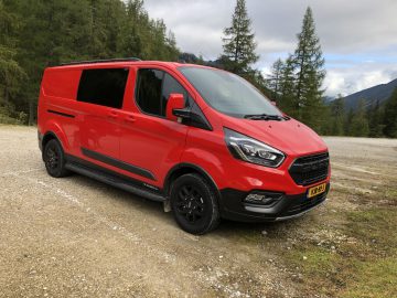 Een rode Ford Transit Custom Trail met zwarte accenten staat geparkeerd op een onverharde weg in een bosrijk bergachtig gebied onder een gedeeltelijk bewolkte hemel.