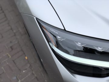 Close-up van de linkervoorhoek van een zilveren Lexus LS-auto, waarbij de nadruk ligt op de koplamp en een deel van het spatbord, met een geplaveide straat zichtbaar op de achtergrond.