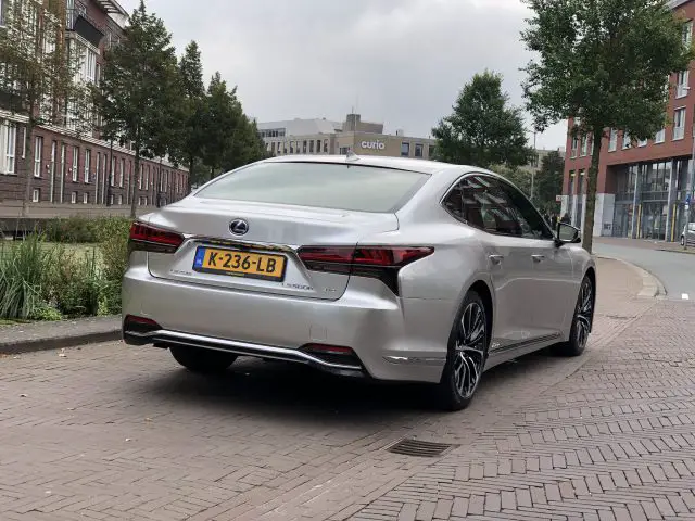 Een zilverkleurige Lexus LS sedan met Nederlands kenteken K-236-LB staat geparkeerd in een geplaveide straat met bomen en moderne gebouwen op de achtergrond.