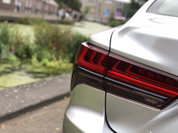 Close-up van het achterlicht rechtsachter van een zilverkleurige Lexus LS geparkeerd nabij een groen gebied met planten en een pad zichtbaar op de achtergrond.