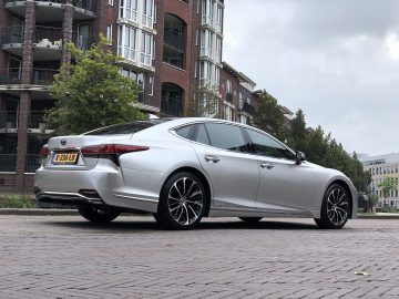 Een zilveren Lexus LS sedan staat geparkeerd in een geplaveide straat in een stadsgebied met moderne appartementsgebouwen en bomen op de achtergrond. Het kenteken van de auto is zichtbaar.