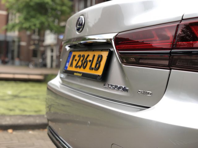 Close-up van de achterkant van een zilveren Lexus LS met een zichtbaar kenteken met de tekst "XT-236-LB". De auto staat buiten geparkeerd in de buurt van een groene vijver.