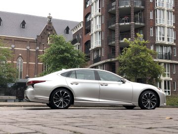 Een zilverkleurige Lexus LS sedan staat geparkeerd voor een kerk en een modern appartementencomplex, met bomen er vlakbij.