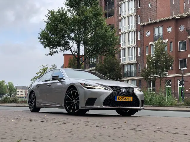 Een zilveren Lexus LS staat geparkeerd in een straat voor een bakstenen woongebouw met meerdere verdiepingen, met bomen en andere gebouwen zichtbaar op de achtergrond.