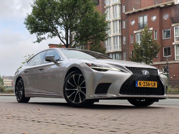 Een zilverkleurige Lexus LS sedan geparkeerd in een bakstenen straat, met een appartementengebouw en bomen op de achtergrond. Op het kenteken staat "K-236-LB".