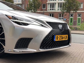 Close-up van de voorkant van een zilveren Lexus LS met Nederlands kenteken "K-236-LB", geparkeerd voor een bakstenen woongebouw.