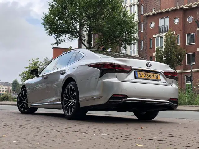 Een zilverkleurige Lexus LS sedan staat geparkeerd in een geplaveide straat voor een bakstenen gebouw, met op de achtergrond bomen en andere woongebouwen zichtbaar.