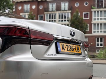 Close-up van de achterkant van een zilverkleurige Lexus LS 500h geparkeerd in een straat, met een gele kentekenplaat met "K-236-LB" voor een rode bakstenen appartementencomplex met meerdere ramen.