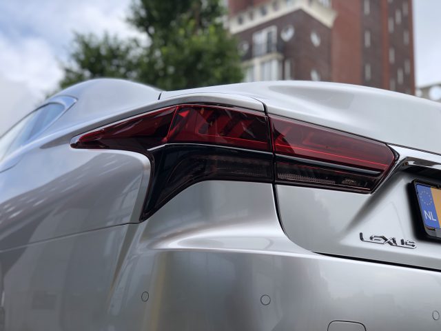 Close-up van het achterlicht linksachter en het embleem van een zilverkleurige Lexus LS die buiten geparkeerd staat, met op de achtergrond een bakstenen gebouw en bomen.