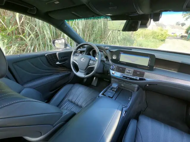 Binnenaanzicht van een moderne Lexus LS met het stuur, het dashboard en het touchscreen. De auto is voorzien van zwart lederen stoelen en diverse bedieningsknoppen op de console. Door de ramen is het groen zichtbaar.