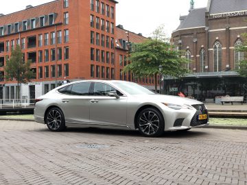 Een slanke luxe sedan Lexus LS staat geparkeerd op een geplaveide weg in een stedelijk gebied, omlijst door bakstenen gebouwen en een hotel op de achtergrond.