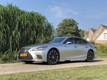 Een zilveren Lexus LS sedan staat geparkeerd op een verhard pad naast hoge groene planten. Onder een helderblauwe lucht zijn op de achtergrond huizen zichtbaar.