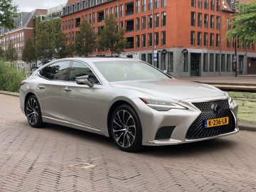 Een slanke zilveren Lexus LS sedan staat geparkeerd in een stadsstraat, omlijst door een rood bakstenen gebouw en weelderige bomen op de achtergrond. Het voertuig is voorzien van een geel kenteken.