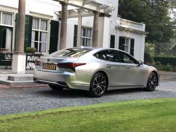 Een zilverkleurige Lexus LS sedan met Nederlands kenteken staat geparkeerd op een grindoprit voor een wit gebouw met groene luiken en kolommen.