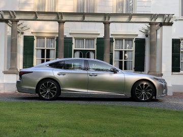 Een zilveren Lexus LS sedan staat geparkeerd voor een wit gebouw met groene luiken en kolommen, omgeven door een weelderig gazon.