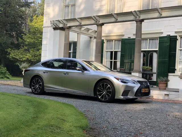 Een zilveren Lexus LS sedan staat geparkeerd op een grindoprit voor een wit huis van twee verdiepingen met groene luiken.