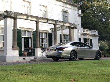 Een zilverkleurige Lexus LS sedan staat geparkeerd voor een groot wit huis van twee verdiepingen met kolommen en groene luiken. De oprit is van grind, met een goed onderhouden gazon op de voorgrond.