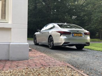 Een zilveren Lexus LS sedan staat geparkeerd op een grindoprit naast een wit gebouw, omgeven door groen. Het kenteken van de auto is zichtbaar.