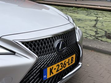 Een close-up van de grille van een zilveren Lexus LS-auto met een geel Nederlands kenteken met de tekst 'K-236-LB'. De auto staat geparkeerd bij een kanaal met met algen bedekt water.