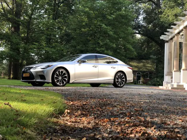 Een zilveren Lexus LS staat geparkeerd op een grindoprit naast een wit gebouw en weelderige groene bomen.