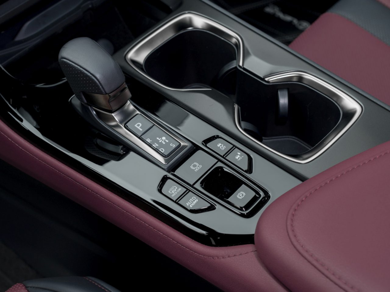 Close-up van de middenconsole van een Lexus NX met een versnellingspook, meerdere bedieningsknoppen en bekerhouders. Het interieur bestaat uit een kastanjebruin en zwart lederen ontwerp.