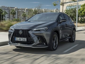 Een slanke grijze Lexus NX SUV rijdt door een drukke stadsstraat, met torenhoge gebouwen en weelderige bomen op de achtergrond.