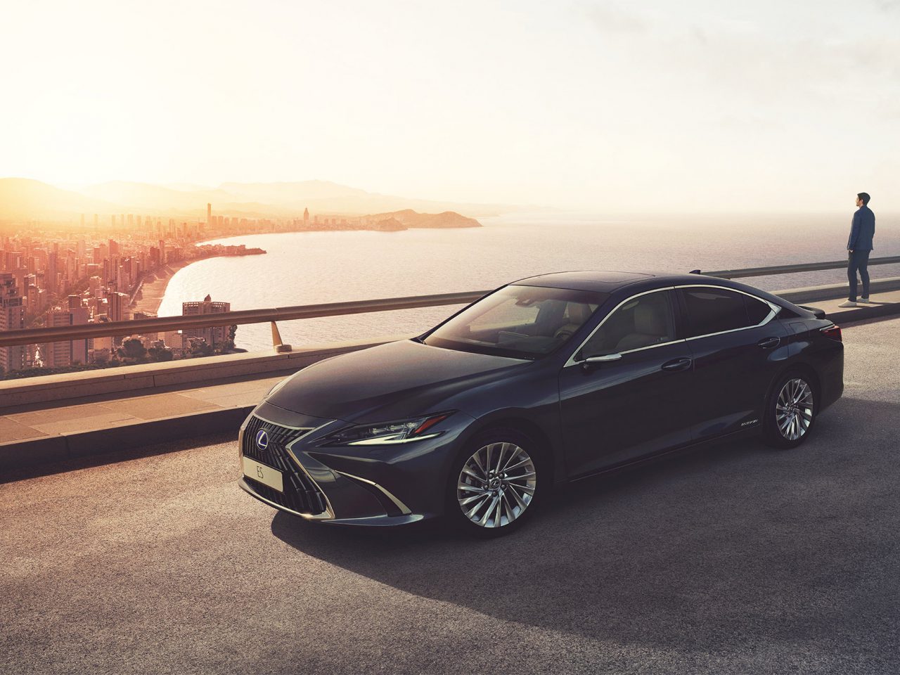 Een strakke zwarte Lexus geparkeerd op een weg in de buurt van een watermassa met een stadsgezicht op de achtergrond bij zonsondergang; een persoon staat op de rand en bewondert het uitzicht.