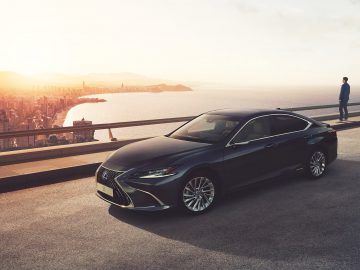 Een strakke zwarte Lexus geparkeerd op een weg in de buurt van een watermassa met een stadsgezicht op de achtergrond bij zonsondergang; een persoon staat op de rand en bewondert het uitzicht.
