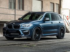 In een straat vlakbij een industrieel gebouw staat een matblauwe BMW X3 SUV geparkeerd, met op maat gemaakte zwarte velgen en een sportief design.