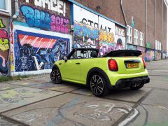 Een felgele MINI Cabrio staat geparkeerd voor een kleurrijke, met graffiti bedekte muur. De kap van de auto is naar beneden en de scène lijkt zich in een stedelijke omgeving af te spelen.