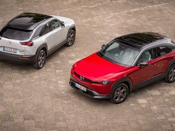 Twee Mazda MX-30 crossover-SUV's geparkeerd op de stoep: de ene is wit en de andere rood, beide met zwarte daken en zilverkleurige lichtmetalen velgen.