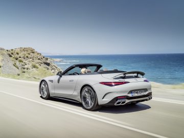 Een witte Mercedes-AMG SL cabrioletsportwagen die onder een heldere hemel over een kustweg rijdt met de oceaan op de achtergrond.
