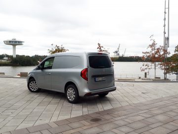 Een zilveren Mercedes-Benz Citan-busje staat geparkeerd op een verhard terrein naast een watermassa. Op de achtergrond zijn er bomen, een kraan en een witte torenconstructie. Op het kenteken staat S-KC 1004.