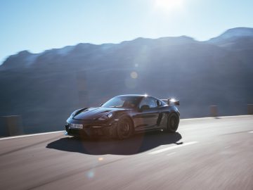 Een Porsche 718 snelt over een bergachtige weg onder een felle zon, met vage bergtoppen op de achtergrond.