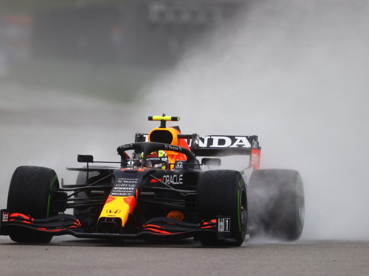 Een Formule 1-auto met Honda-logo, bestuurd door Max Verstappen, snelt op een natte baan, waardoor er een waterstraal achter ontstaat. De bestuurder draagt een felgeel-oranje helm.