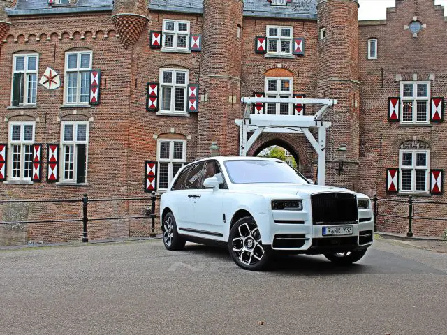 Een witte Rolls-Royce Cullinan Black Badge staat geparkeerd voor een historisch bakstenen kasteel met decoratieve ramen en een kleine ophaalbrug.
