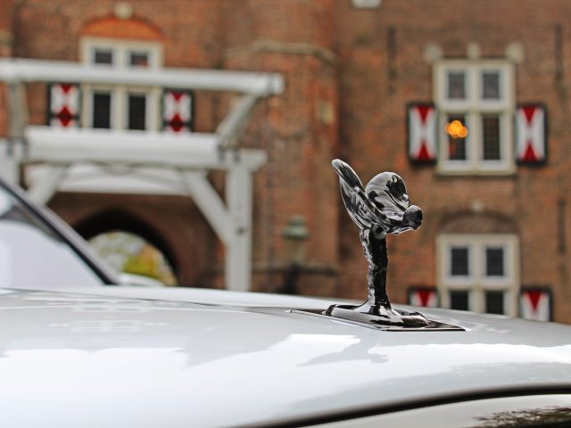 Close-up van een ornament op de motorkap van een auto in de vorm van een vliegend figuur op een Rolls-Royce Cullinan Black Badge, met een onscherpe achtergrond die een bakstenen gebouw laat zien met ramen met witte kozijnen en een decoratieve boog.