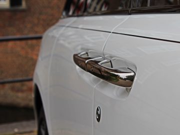 Close-up van de zijkant van een Rolls-Royce Cullinan Black Badge, gericht op de chromen deurgrepen, met een wazig bakstenen gebouw op de achtergrond.